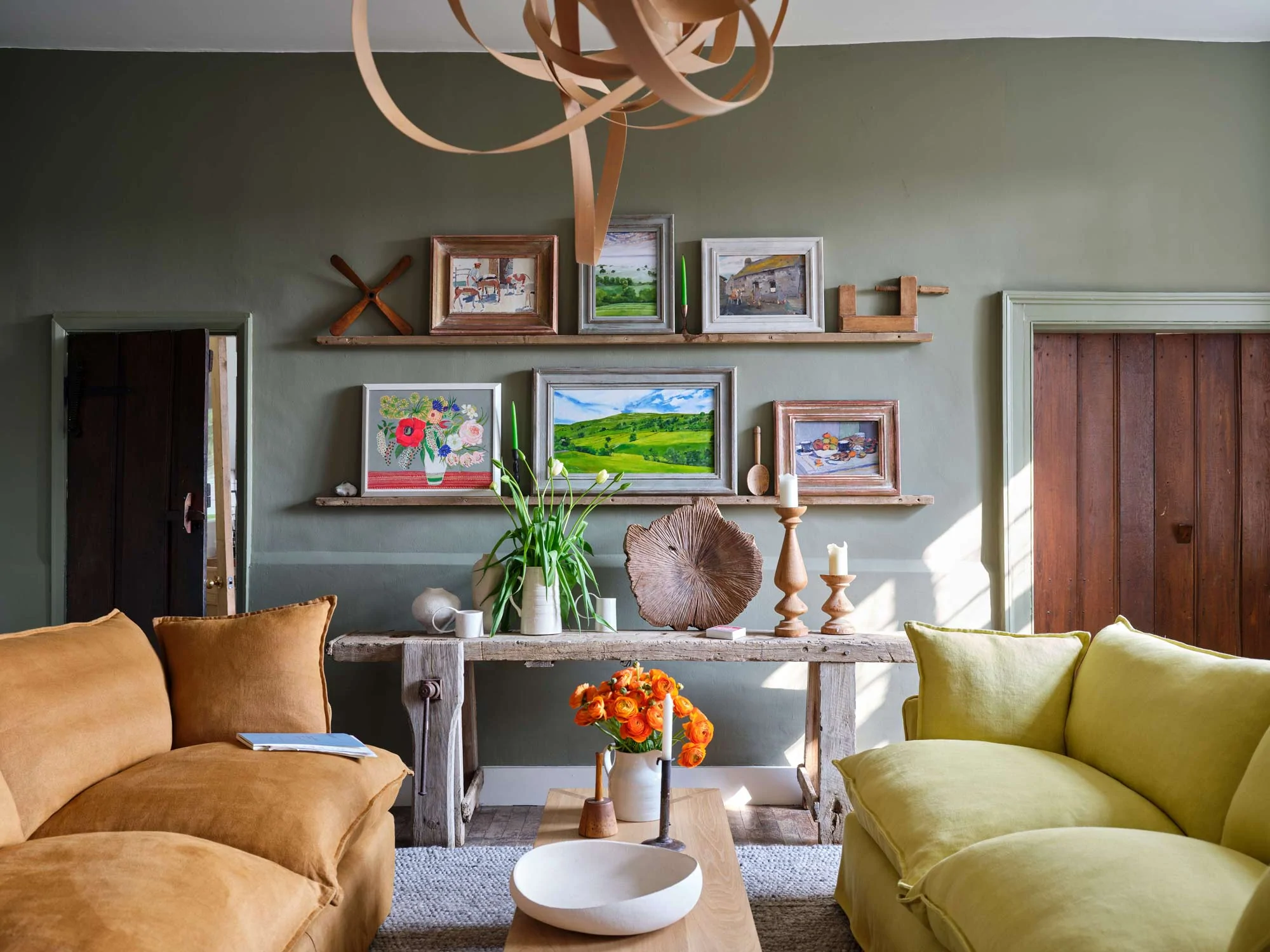 A soft warm moss green room with a citrine yellow and brown bronzite sofa facing each other with a birch coffee table in between The walls are covered in paintings and decorations in 3 horizontal rows Against the wall is a rustic wooden side table with candles a vase of white tulips and a wooden sculptural bowl
